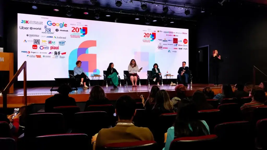 Estive no Congresso da Abraji 2025 e saí com a cabeça fervilhando de ideias 5 Painel de conferência com cinco pessoas sentadas no palco e um apresentador falando ao microfone durante evento em um auditório, no congresso da abraji 2025